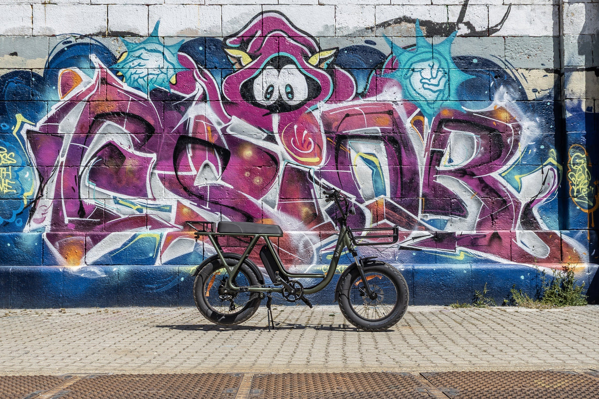 Vélo électrique vert foncé devant un mur de briques orné d'un graffiti coloré violet et bleu.