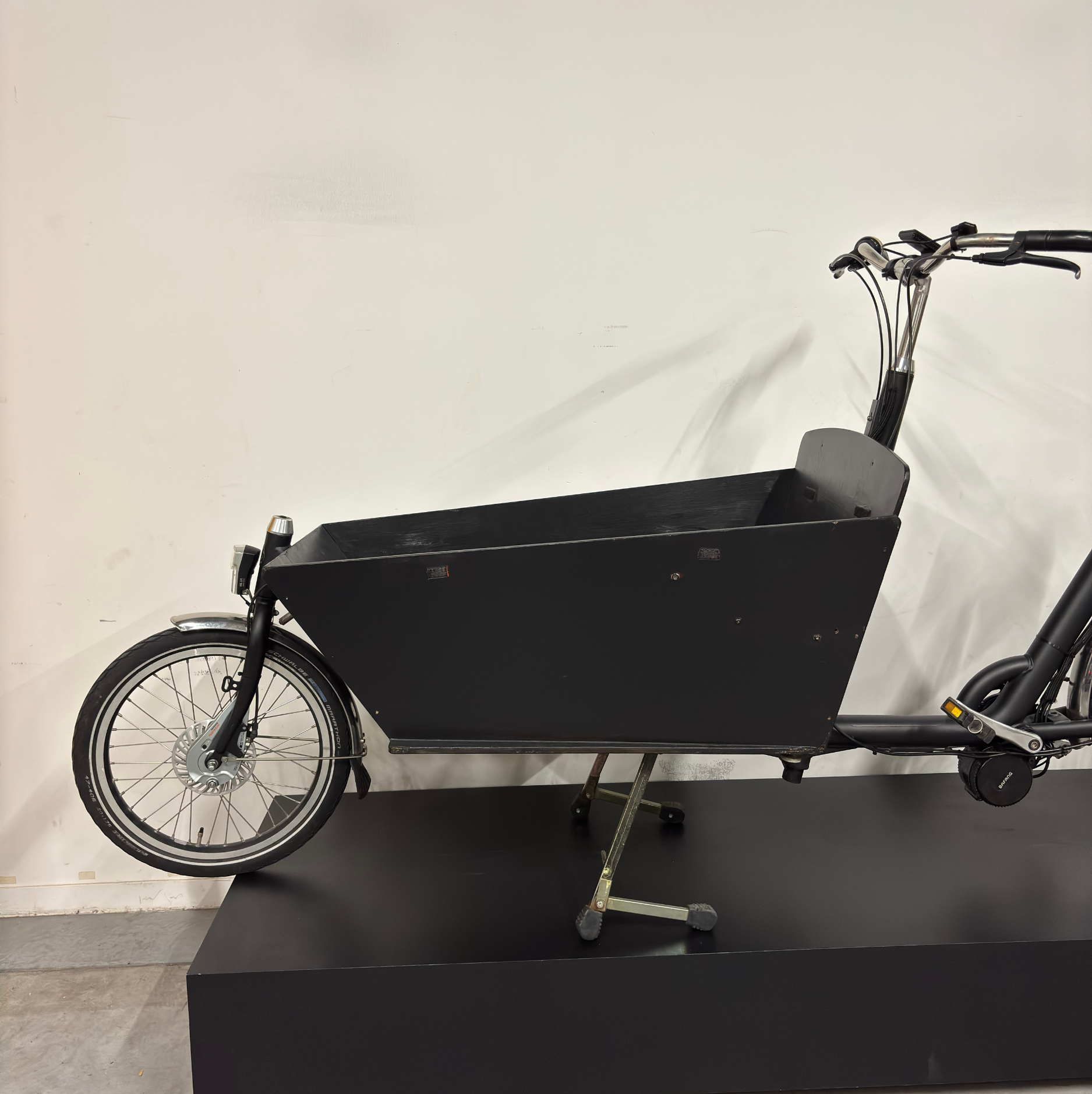 Vue avant du vélo cargo électrique noir WAP Lille, cadrage sur la caisse avant et la petite roue avant posées sur un présentoir noir