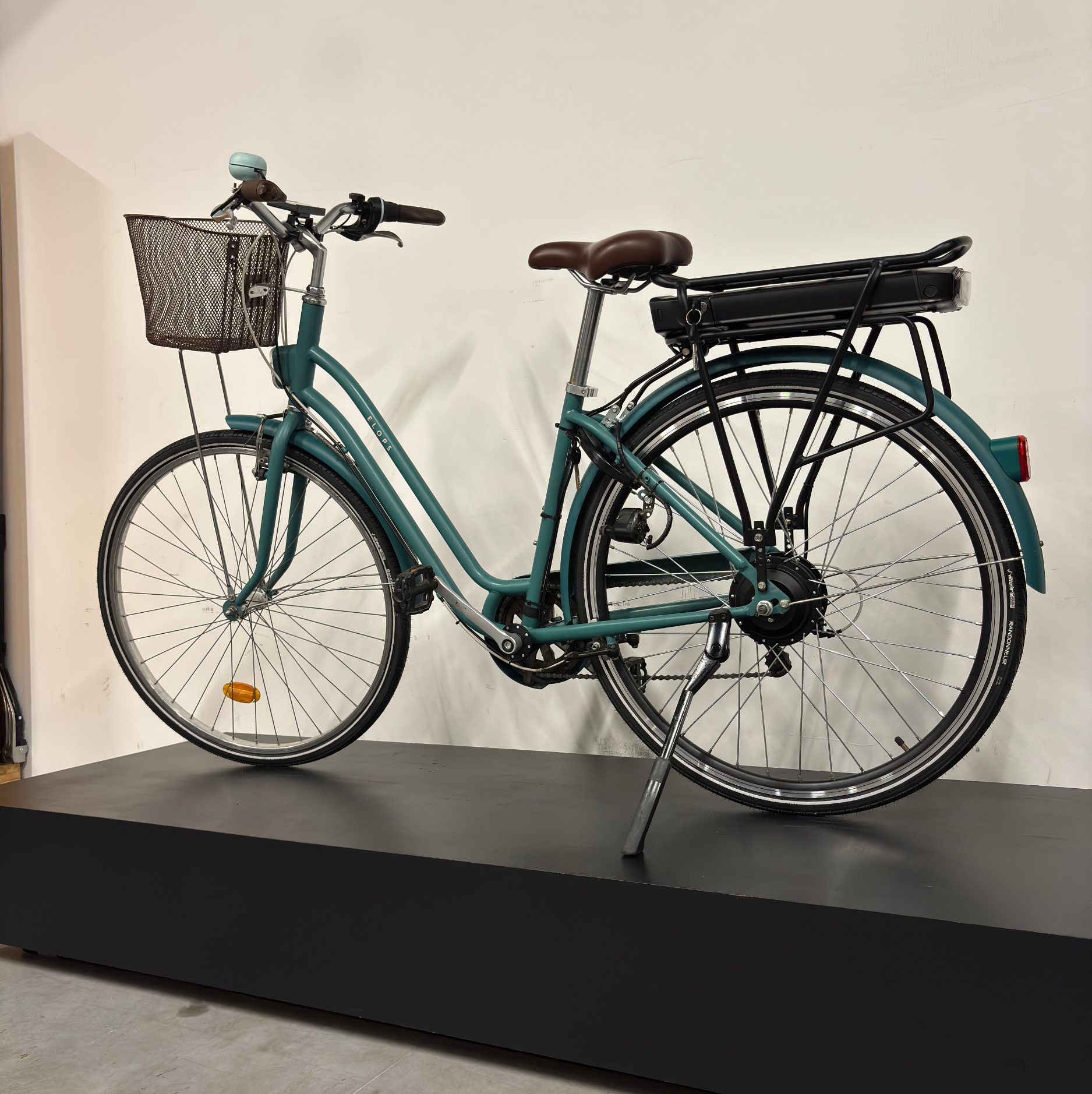 Vélo de ville électrique vert cadre bas WAP Lille, vu de profil côté droit sur présentoir noir, avec panier avant, selle marron et batterie sur porte-bagages