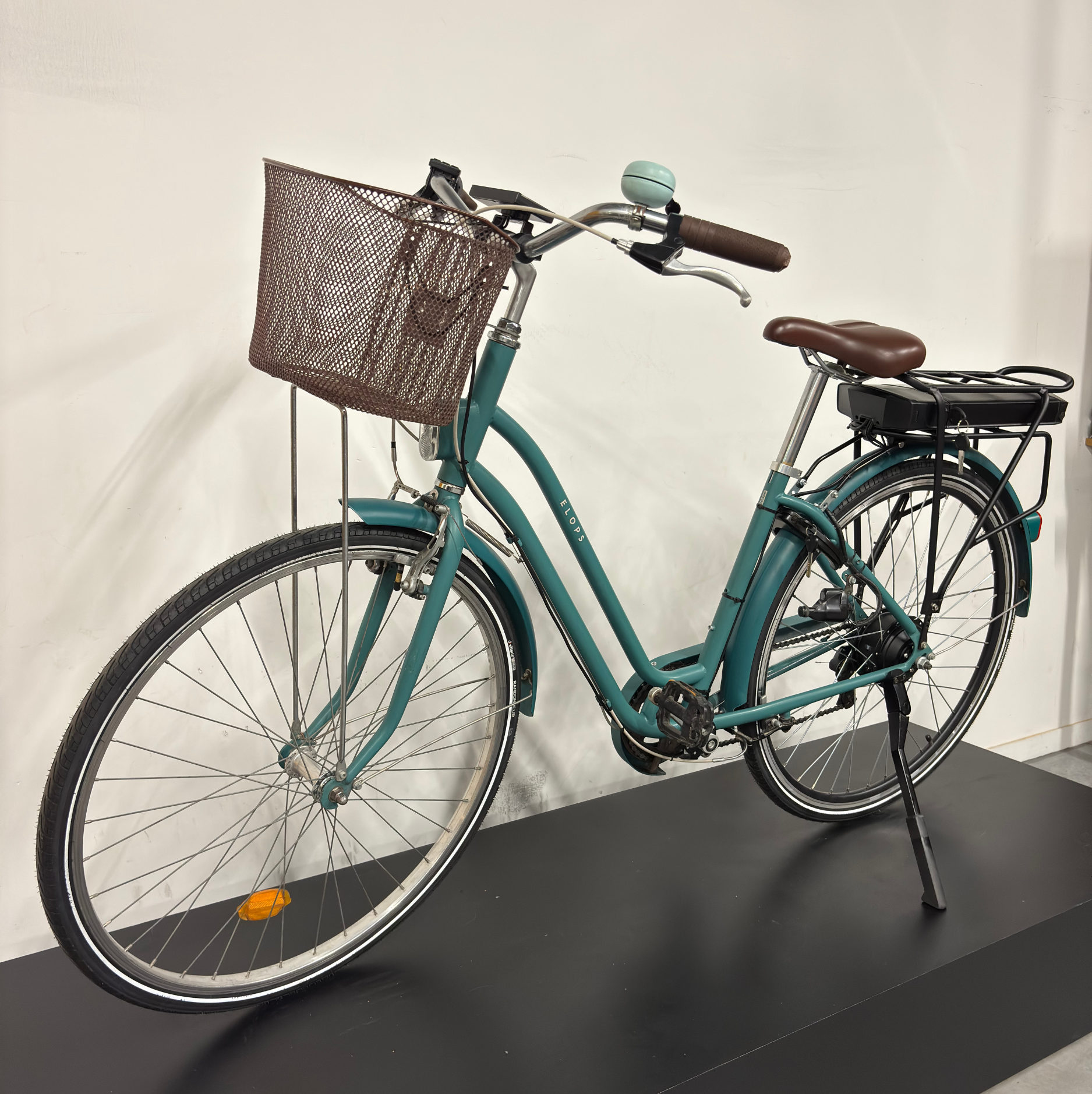 Vélo de ville électrique vert cadre bas WAP Lille, vu de trois quarts avant droit sur présentoir noir, avec panier avant, selle marron et batterie sur porte-bagages